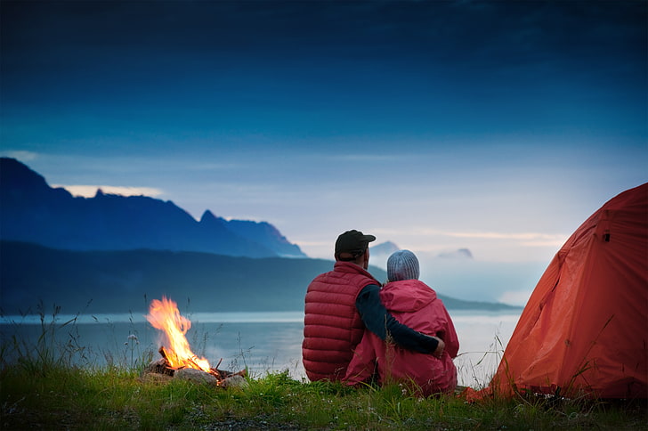 Two campers next to a tent and campire, looking out over the ocean with mountains in the distance
